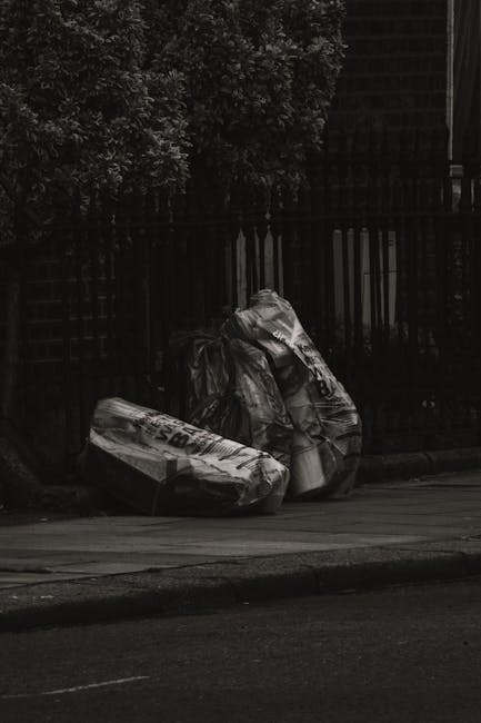 Two large rubbish bags made of black plastic are placed on the pavement near a dark wooden fence, with a backdrop of dense foliage from a bush or tree. The bags appear to contain household waste or di