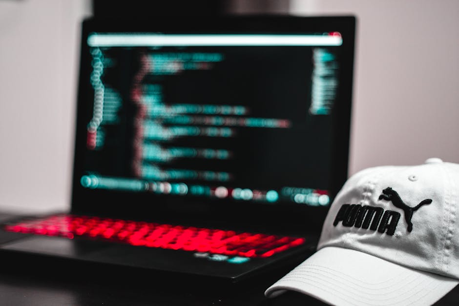A black laptop with a red backlit keyboard displaying a blurred screen of lines of code or scripting text, positioned on a flat surface. To the right of the laptop, a white Puma baseball cap with a bl
